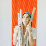 Woman holds three rolls of wallpaper. On background visible wall with orange wallpaper.